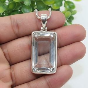 Peut inclure: Pendentif rectangulaire en cristal transparent avec un cadre et une chaîne en métal argenté. Le cristal est facetté, captant la lumière. Le pendentif est tenu dans une main sur un fond de feuillage vert.