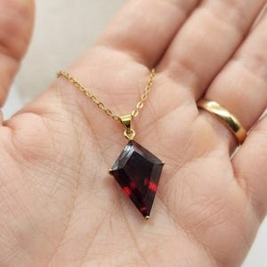May include: A gold chain necklace featuring a kite-shaped, deep red gemstone pendant. The faceted stone is set in a simple gold metal frame, suspended from a delicate gold-toned link chain that rests against the palm of a hand.