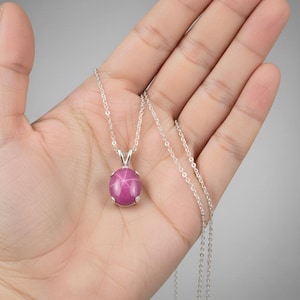Puede incluir: Un collar de plata con un colgante. El colgante presenta una gran gema de rubí estrella ovalada. El rubí es de color rosa intenso y está engastado en un bisel de plata. La cadena es delicada y cuelga del colgante.