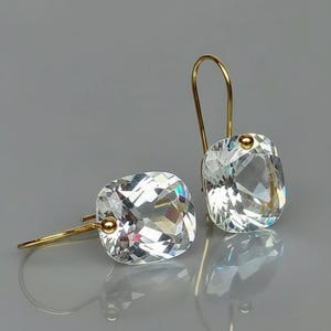 May include: A pair of clear, square-cut crystal earrings with gold-coloured hooks. The crystals are faceted, reflecting light. The earrings are displayed on a reflective surface.