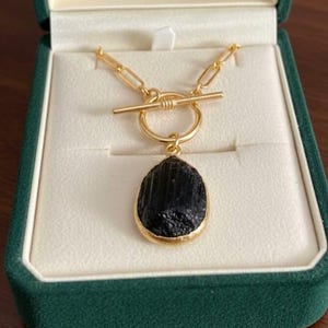 Puede incluir: Un collar dorado con un colgante negro en forma de lágrima. El colgante está engastado en un bisel dorado y cuelga de una cadena dorada con un cierre de palanca. El collar se muestra en una caja de joyería de terciopelo verde.