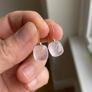 Puede incluir: Un par de pendientes de cuarzo rosa con ganchos dorados. Cada pendiente presenta una piedra de cuarzo rosa facetada en forma cuadrada. Los pendientes son sostenidos por una mano, mostrando el diseño delicado y el tono rosa suave de las piedras preciosas.