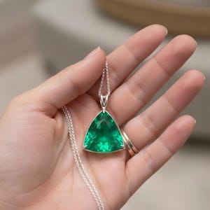 Peut inclure: Un collier en argent avec un pendentif triangulaire en pierre précieuse vert émeraude facettée. Le pendentif est serti dans un cadre argenté et suspendu à une fine chaîne en argent. La pierre a une couleur verte vibrante.