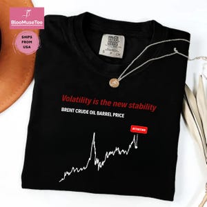 May include: Black t-shirt with the text "Volatility is the new stability" and "BRENT CRUDE OIL BARREL PRICE" in red. A white line graph and the word "ATTENTION" in a red box are also on the shirt. A silver necklace with a pendant is on the shirt.