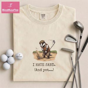 May include: Cream-colored t-shirt with a badger golfer graphic and the text "I HATE SAND. (And yet...)". Includes golf balls and golf clubs. The shirt is made of a soft, comfortable material.