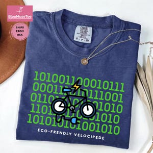 May include: A navy blue t-shirt with a green binary code design and a graphic of an electric bicycle. The text "ECO-FRENDLY VELOCIPEDE" is printed below the bicycle graphic. The shirt has a crew neck and short sleeves.