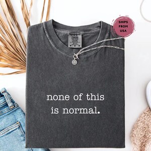 May include: Dark gray Comfort Colors t-shirt with the phrase "none of this is normal" in white. A silver necklace with a circular pendant rests on the shirt. The shirt is folded neatly.