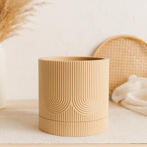 Peut inclure: Un récipient cylindrique beige avec des lignes verticales et des motifs en arc. Le récipient est posé sur une surface claire, avec un plateau tissé et du tissu en arrière-plan. Le design est minimaliste et moderne.
