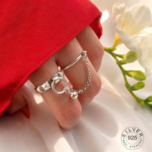 Peut inclure: Une bague en argent sterling composée d'un anneau large à texture cloutée et d'un anneau plus fin reliés par une fine chaîne. La chaîne comporte une petite breloque sphérique pendante. Le métal porte le poinçon 925 pour l'argent sterling.
