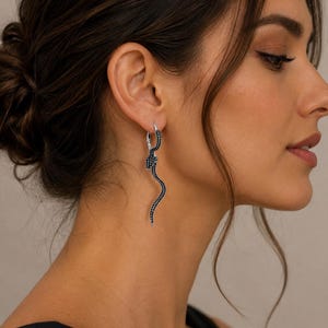 Op de afbeelding: Een zilverkleurige oorring met een bungelende, S-vormige slangen hanger. Het slangenlichaam is ingelegd met talloze kleine, donkerzwarte steentjes die het gehele oppervlak bedekken, wat zorgt voor een getextureerd en glinsterend uiterlijk op het metaal.
