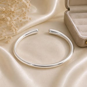 May include: A polished silver-tone cuff bracelet with a smooth, rounded, open-ended design. The metal surface is reflective and features a simple, continuous circular shape with tapered ends, showcasing a clean and uniform metallic finish throughout.