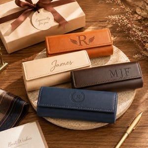Könnte beinhalten: Vier rechteckige Lederetuis mit Klappverschluss, jedes mit individueller Personalisierung: ein hellbraunes Etui mit einem R und Flügeln, ein beiges Etui mit dem Namen James, ein dunkelbraunes Etui mit den Initialen MJF und ein marineblaues Etui mit einem B und Benjamin.
