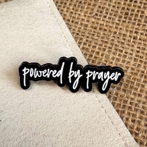May include: A black pin with white cursive lettering that reads "powered by prayer". The pin has a glossy finish and a distinct outline that follows the shape of the text, creating a bold contrast between the black background and white letters.