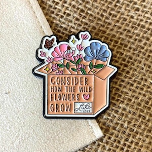 May include: Enamel pin depicting a box overflowing with pink and blue flowers, a butterfly, and the text "Consider how the wild flowers grow." The pin has a black outline and a Luke 12:27 reference.