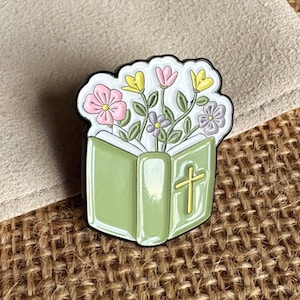 May include: A metal enamel pin shaped like an open light green book with a yellow cross on the cover. A bouquet of small pink, yellow, and purple flowers with green stems and leaves emerges from the top of the book, all outlined in black metal.