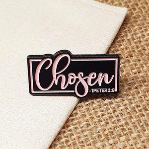 Puede incluir: Un pin de esmalte negro y rosa con la palabra "Chosen" en cursiva, enmarcada por un rectángulo negro. El texto "-1PETER 2:9" está debajo. El pin está sobre una tela blanca y una superficie de yute.