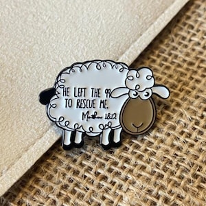 May include: A sheep-shaped enamel pin with a white body, black accents, and a brown face. The pin features the text "He left the 99 to rescue me. Matthew 18:12" in black lettering. The pin is set against a neutral background.