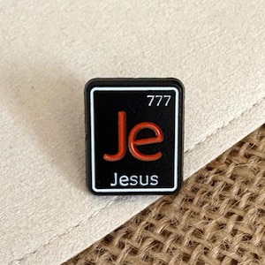 Puede incluir: Un pin de esmalte rectangular con fondo negro y borde blanco. El diseño imita un elemento de la tabla periódica con las letras grandes de color naranja Je, el número 777 en la esquina superior derecha y la palabra Jesus impresa debajo.