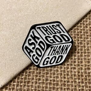 Puede incluir: Un pin de esmalte negro y blanco en forma hexagonal. El pin presenta las palabras "ASK GOD", "TRUST GOD" y "THANK GOD" en una fuente en negrita sin serifa. El pin está sobre un fondo neutro.