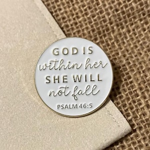 May include: A white and gold enamel pin with the text "GOD IS within her SHE WILL not fall" and "PSALM 46:5". The pin is round and has a gold border. The pin is on a beige surface.