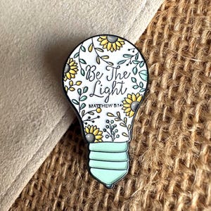 May include: A lightbulb-shaped enamel pin featuring a white background with yellow sunflowers, green leaves, and black vines. The text reads Be The Light and MATTHEW 5:14 in black script and print. The base of the bulb is a solid light mint green color.