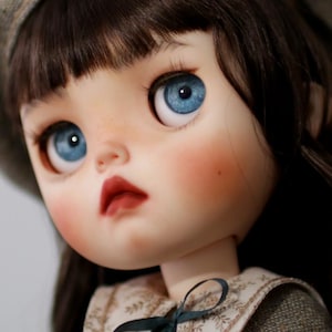 May include: A detailed image of a doll's face, featuring striking blue eyes, flushed cheeks, and dark brown hair. The doll is wearing a grey hat and a dress with a floral collar and a dark green bow. The doll's lips are a deep red.