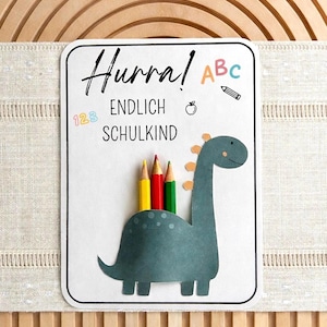 Puede incluir: Una tarjeta blanca con borde negro presenta un dinosaurio verde azulado que sostiene lápices de colores. La tarjeta dice "Hurra! Endlich Schulkind" con ABC, un lápiz, una manzana y los números 123. El fondo es un arco de madera.