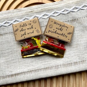 May include: Two small cellophane bags of gummy candies with German text on small brown cards. The bags are labeled "Goldb&auml;re" and contain colorful gummy bears and fruit shapes. The cards have smiley faces and read "Falls es ohne mich z&auml;h wird" and "Wenigstens nicht geschmacklos."