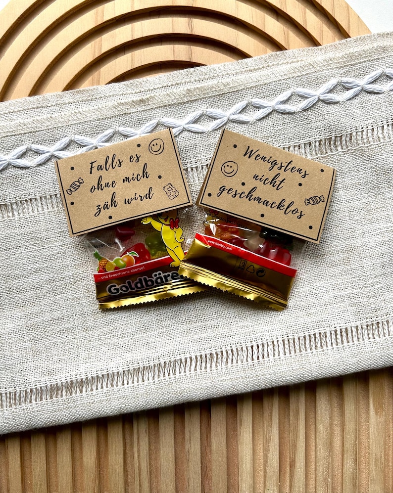 May include: Two small cellophane bags of gummy candies with German text on small brown cards. The bags are labeled "Goldb&auml;re" and contain colorful gummy bears and fruit shapes. The cards have smiley faces and read "Falls es ohne mich z&auml;h wird" and "Wenigstens nicht geschmacklos."