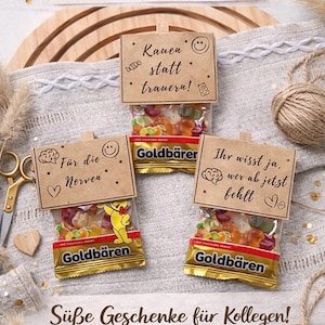 May include: Three small bags of gummy candies with attached tags. The tags have handwritten text and small illustrations. The bags are labeled "Goldb&auml;ren". The image has text in German that says "Sweet gifts for colleagues!"
