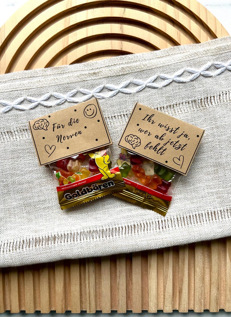 May include: Two small bags of Goldb&auml;ren gummy candies with brown paper toppers. One topper reads "F&uuml;r die Nerven" with a brain graphic, a heart, and a smiley face. The other reads "Ihr wisst ja, wer ab jetzt fehlt" with a brain graphic and a heart.