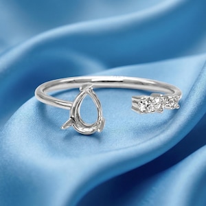 May include: A silver-toned open ring featuring a pear-shaped setting on one end and three small, clear, round-cut stones set in a row on the other end. The band is smooth and polished, creating a delicate and refined appearance for this jewelry piece.