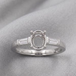 May include: A silver-toned metal ring featuring a central four-prong setting designed for an oval-shaped stone. The band includes a single baguette-cut clear stone on each side of the center setting, creating a three-stone style ring design.
