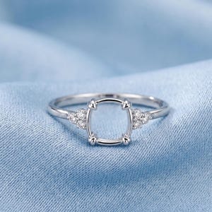 May include: A silver-toned ring featuring a central empty four-prong setting designed to hold a gemstone. The band is accented on each side by a cluster of three small, clear, round-cut stones embedded into the metal shank of the ring.