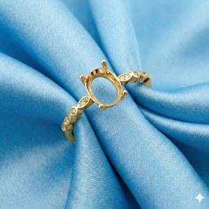 May include: A gold ring setting with an oval center and small prongs. The band features alternating small clear stones and decorative gold accents. The ring is displayed on a blue satin fabric.