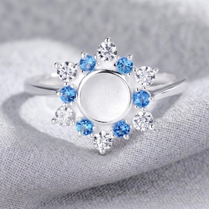 May include: A silver-toned ring featuring a central circular setting surrounded by an alternating pattern of clear white and bright blue round-cut stones. The arrangement of the stones creates a star-like or floral shape around the empty center.
