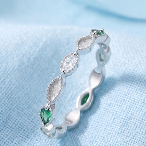 Könnte beinhalten: Ein silberfarbener Ring mit einer Reihe abwechselnder marquiseförmiger Fassungen. Das Design umfasst klare weiße Steine und leuchtend grüne Steine, die in texturierte, gewellte Metallrahmen eingefasst sind und ein durchgehendes Muster bilden.