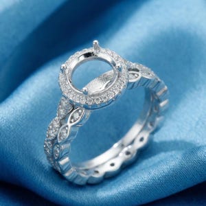 May include: A silver-toned ring setting featuring a circular center mount designed for a gemstone, surrounded by a halo of small clear stones. The band displays an intricate pattern of marquise-shaped links, each embedded with tiny clear stones.