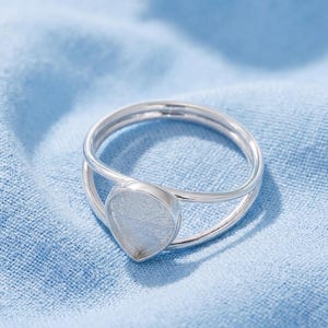 May include: A silver-toned ring featuring a split band design and a teardrop-shaped bezel setting. The metal has a polished finish, and the central setting is empty, designed to hold a stone or inlay. The ring is constructed from a single piece of metal.