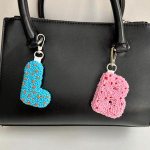 May include: Two handmade keychains shaped like the letters "L" and "B" hang from a black purse. The "L" is light blue with orange dots, and the "B" is pink with red dots. Both have silver-colored clasps.