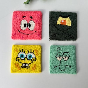 May include: Four square, textured coasters featuring cartoon characters. The coasters include Patrick Star in pink, Plankton in dark green, SpongeBob SquarePants in yellow, and Squidward in light green. Each coaster has a different character's face.