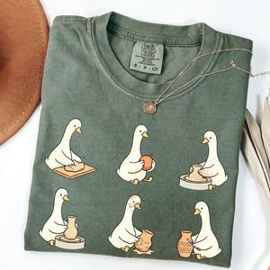 May include: An olive green t-shirt featuring a graphic print of six white geese engaged in various stages of pottery making, including kneading clay, shaping a vessel on a wheel, and painting a finished pot. The shirt has a Comfort Colors brand label.