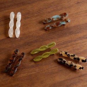 May include: A collection of hair clips and barrettes in various colors and patterns, including white, tortoiseshell, olive green, and a blue-brown mix. The clips are arranged on a wooden surface. The text "Hair Clips & Barrettes" is visible.