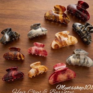 Op de afbeelding: Een verzameling kleine haarclips in verschillende kleuren, waaronder bruin, rood en crème, met gemarmerde patronen. De clips liggen op een houten oppervlak. De tekst "Hair Clips & Barrettes" is zichtbaar.