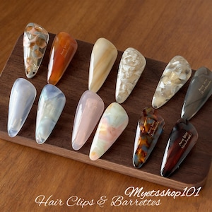 Puede incluir: Una colección de doce pinzas y horquillas decorativas para el cabello en varios colores y patrones. Las pinzas se exhiben en una bandeja de madera. Las pinzas tienen un acabado brillante y son en tonos marrones, beige y colores pastel. El texto "Hair Clips & Barrettes" y "Myetsshop101" son visibles.