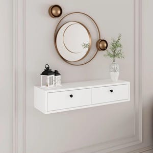 Può includere: Una consolle bianca sospesa a parete con due cassetti e pomelli neri. Sopra, uno specchio rotondo con cornice dorata e dettagli dorati decorativi. Due piccole lanterne bianche e nere e un vaso bianco con vegetazione.