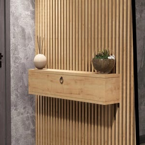 Puede incluir: Una estantería flotante de madera clara con un cajón y un pequeño tirador redondo negro. La estantería está montada en una pared vertical de listones de madera. Un jarrón decorativo y un terrario de cristal con plantas se encuentran en la estantería.
