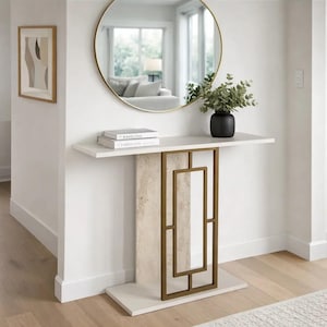 Puede incluir: Una consola blanca con un marco geométrico de metal dorado y una columna central con aspecto de piedra. Un espejo redondo dorado cuelga sobre la mesa, y un jarrón negro con vegetación está sobre la mesa. Dos libros están apilados sobre la mesa.