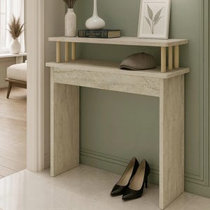 Puede incluir: Una consola de dos niveles de color claro con una parte superior rectangular y un estante inferior. El estante superior contiene jarrones, un libro y una imagen enmarcada. Una gorra marrón descansa sobre el estante inferior. Zapatos de tacón negros se colocan debajo de la mesa.