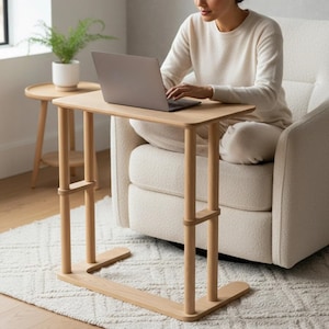 Può includere: Un tavolino regolabile per laptop in legno chiaro, con piano rettangolare e gambe arrotondate. Il tavolo è sostenuto da due montanti verticali con impostazioni di altezza regolabili. Un tavolino e una pianta in vaso sono visibili sullo sfondo.